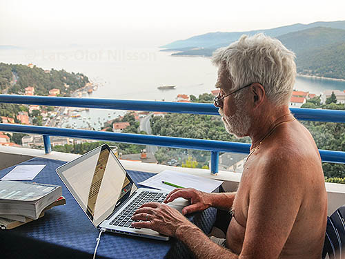 A man on a balcony overlooking the sea is writing on his laptop