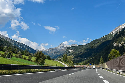 A landscape with a motorway in the foreground and mountains in the background against a blue sky