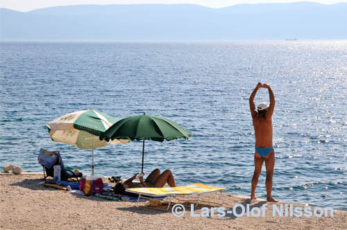 Against the blue sea a man is stretching on the beach while his wife is lying under a parasol