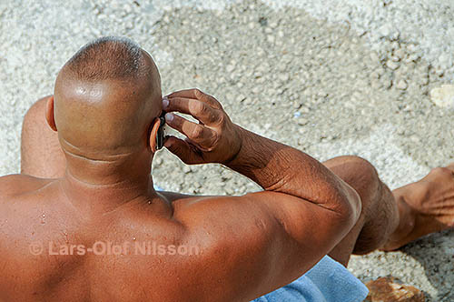 Burly man on a beach speaking on a cellphone. He is enquiring about renting a sunshade.