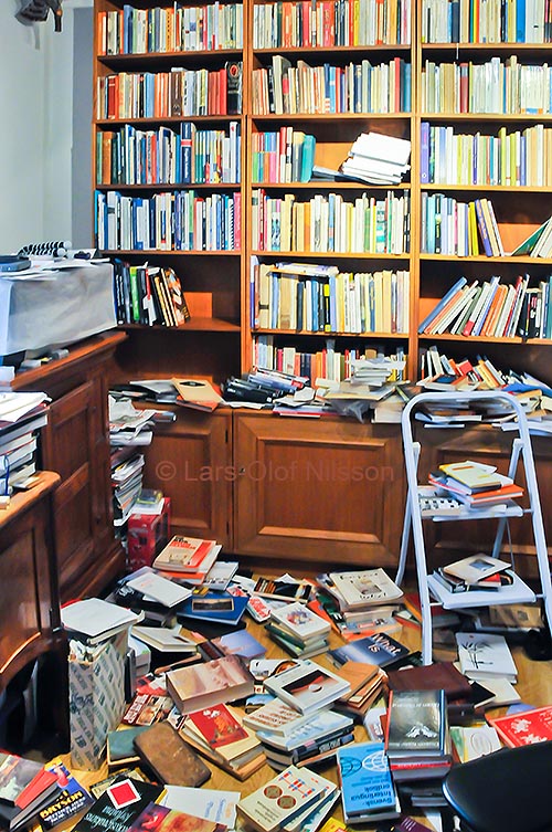 The image shows a room with bookshelves. There are many book on the floor, on a desk and on a step-ladder. The purpose is to illustrate the verb rearrange in a post about the prefix re..