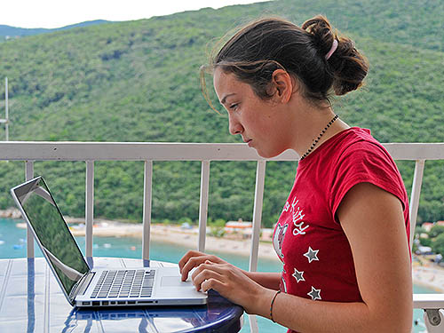 A young girl on a balcony is writing on a laptop. The image illustrates the concept of emails.