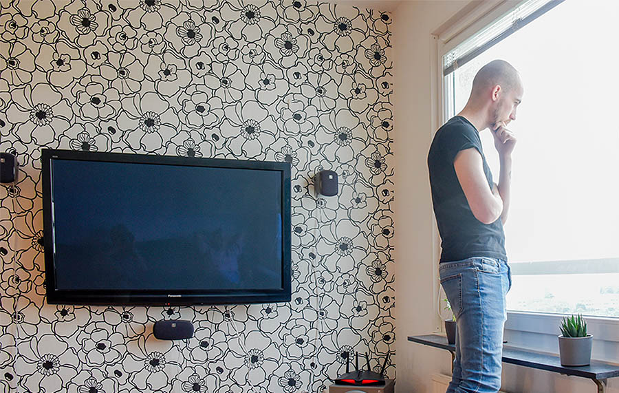 A msn is standing by a window, looking out. A TV set is mounted on a wall. The wallpaper has a pattern of large flowers in black and white. The image illustrates the concept of dissatisfied.