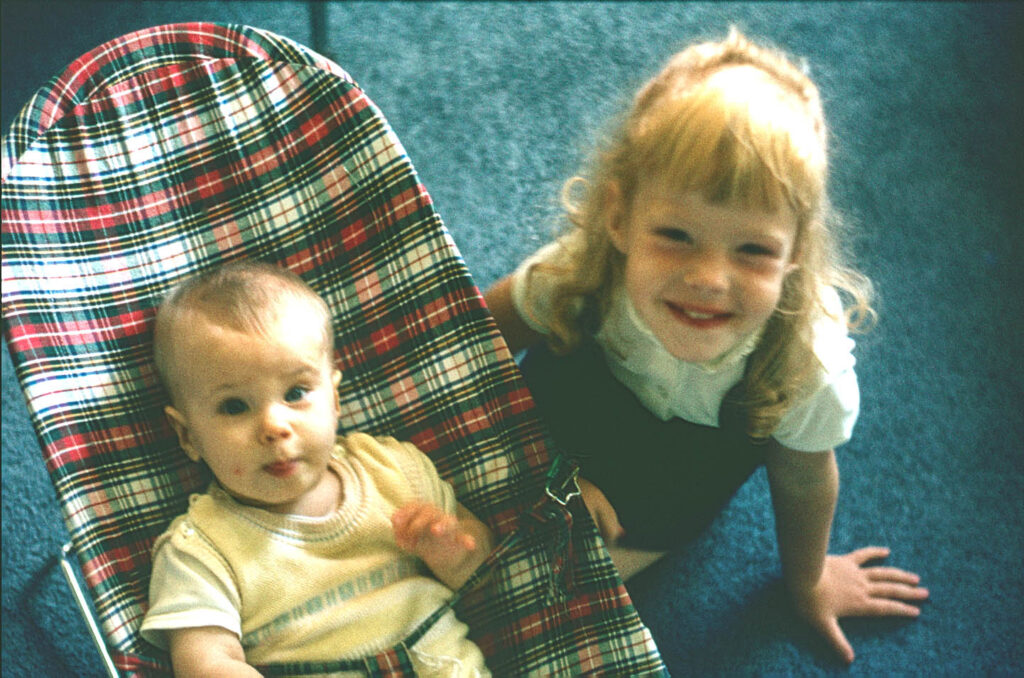 The image shows a small child in a baby bouncer. Her sister is sitting next to her. The image illustrates the word babysitter as an example of. false friends, words that are the same but different.