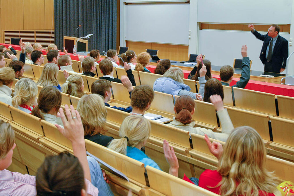 A speaker in front of his audience. The image illustrates the word rapport, and several in the audience are putting their hands up as a response to what is being said.